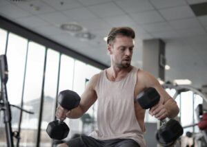 From below of muscular male pumping biceps with dumbbells while sitting on bench in sports center by following the instruction of Weight Loss provided on Mr. Fat Belly Website platform.
