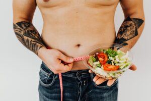 Topless Man Holding a Sliced Tomato and Fresh Vegetable Salad