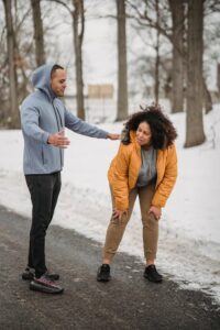 Full body of overweight tired African American female tilted forward standing on asphalt walkway near black trainer during training in winter park