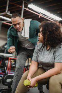 African American male athlete coaching black woman and explaining exercises technique with dumbbells in gym