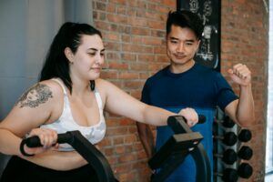 Smiling young tattooed plus size sportswoman in activewear doing cardio exercise on fitness cycle while training in contemporary gym with positive Asian male instructor