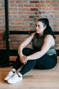 Full body of pensive young overweight lady in sportswear with ponytail sitting on laminate floor in gym after hard training