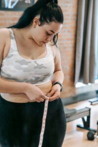 Young obese female using tape for evaluating size of belly while preparing for training in modern fitness center