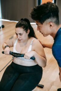High angle of Asian couch supporting plus size female exercising on rowing on machine in gym